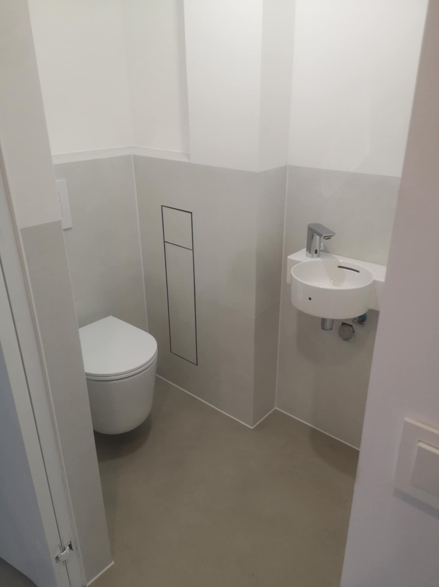 Nowoczesna, minimalistyczna toaleta z podwieszaną miską WC i małą, okrągłą umywalką nablatową z chromowaną baterią, wykończona w odcieniach bieli i szarości z ukrytą instalacją wodną.