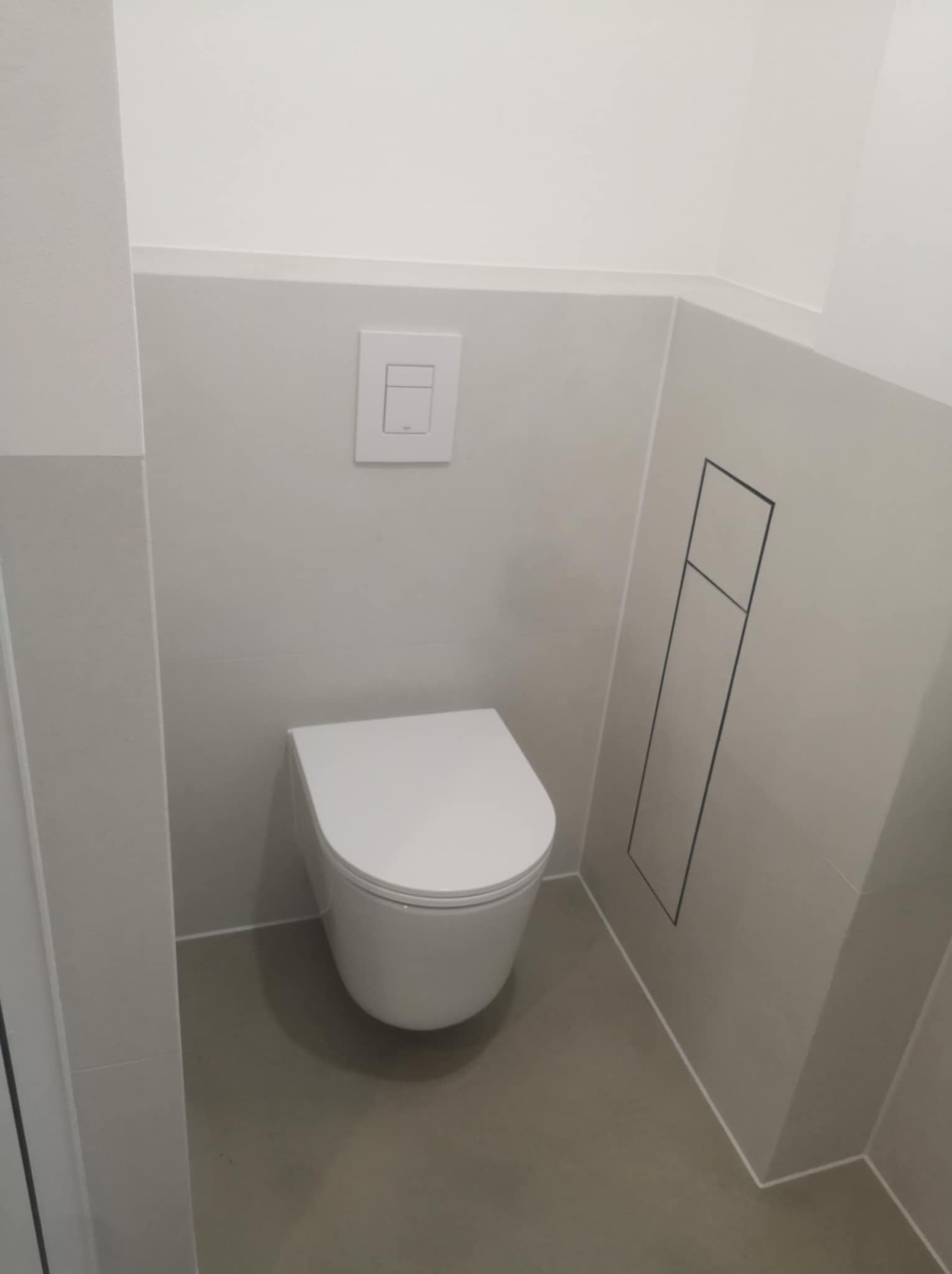 Nowoczesna toaleta podwieszana w minimalistycznej łazience z szarymi płytkami i ukrytym systemem spłukiwania. Na ścianie widoczny biały przycisk spłukujący.