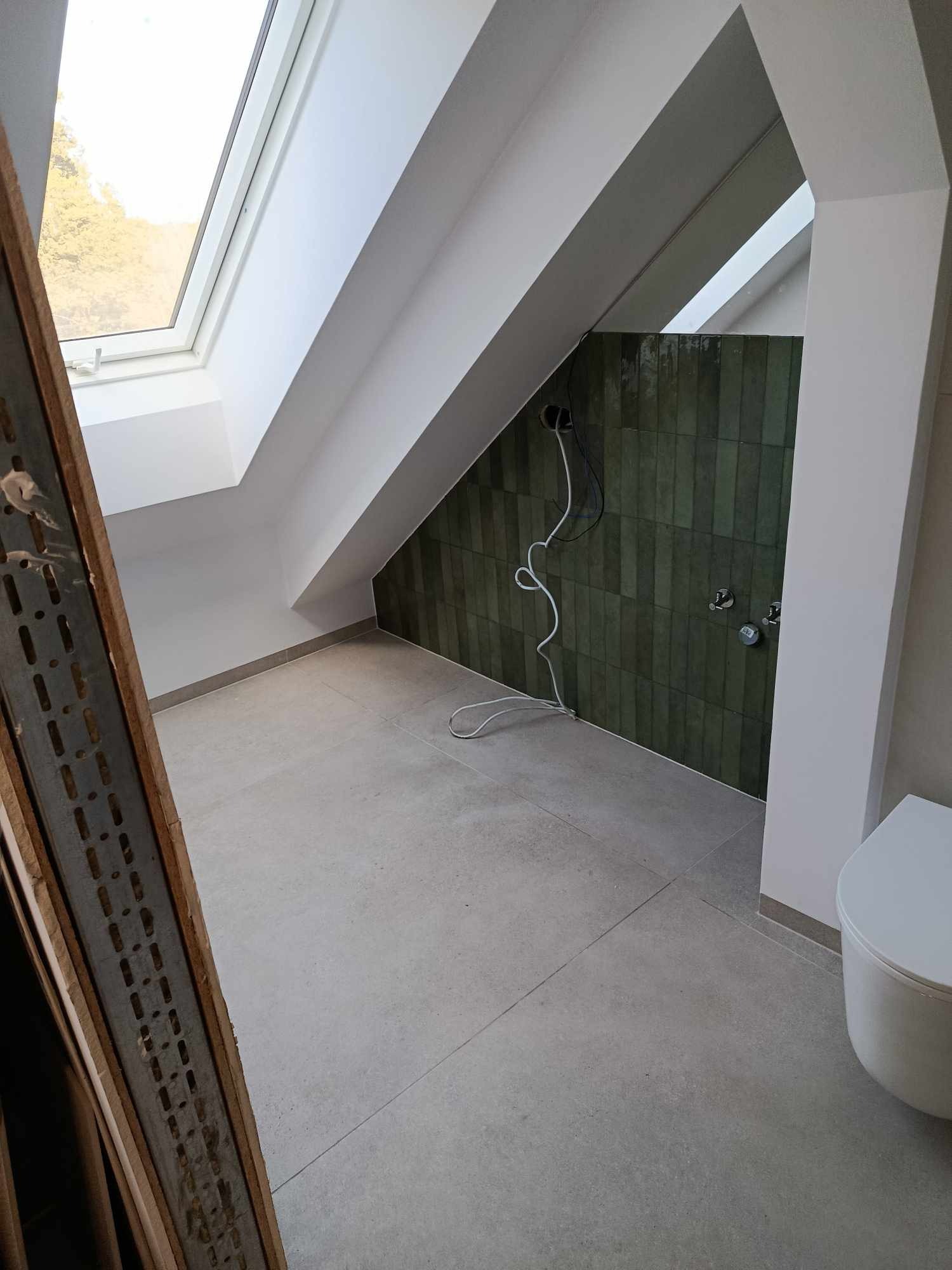 Poddasze z oknem dachowym w trakcie prac wykończeniowych łazienki: ułożone szarobeżowe płytki podłogowe, ściana z pionowych, zielonych płytek, wystające przewody i częściowo widoczna toaleta.