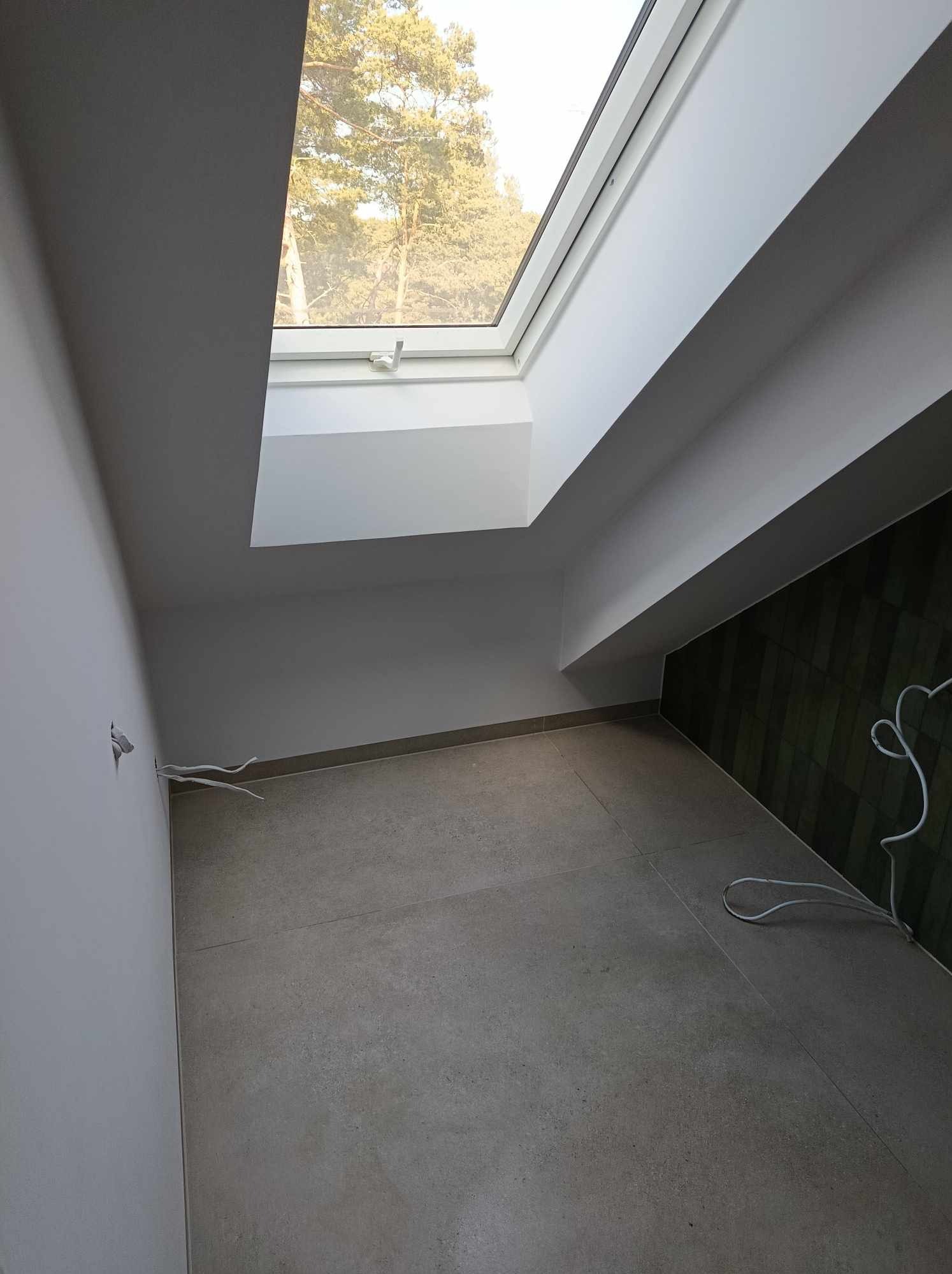 Poddasze z oknem dachowym, szarą podłogą z płytek i fragmentem ściany z zielonych płytek, widoczne wystające kable elektryczne.