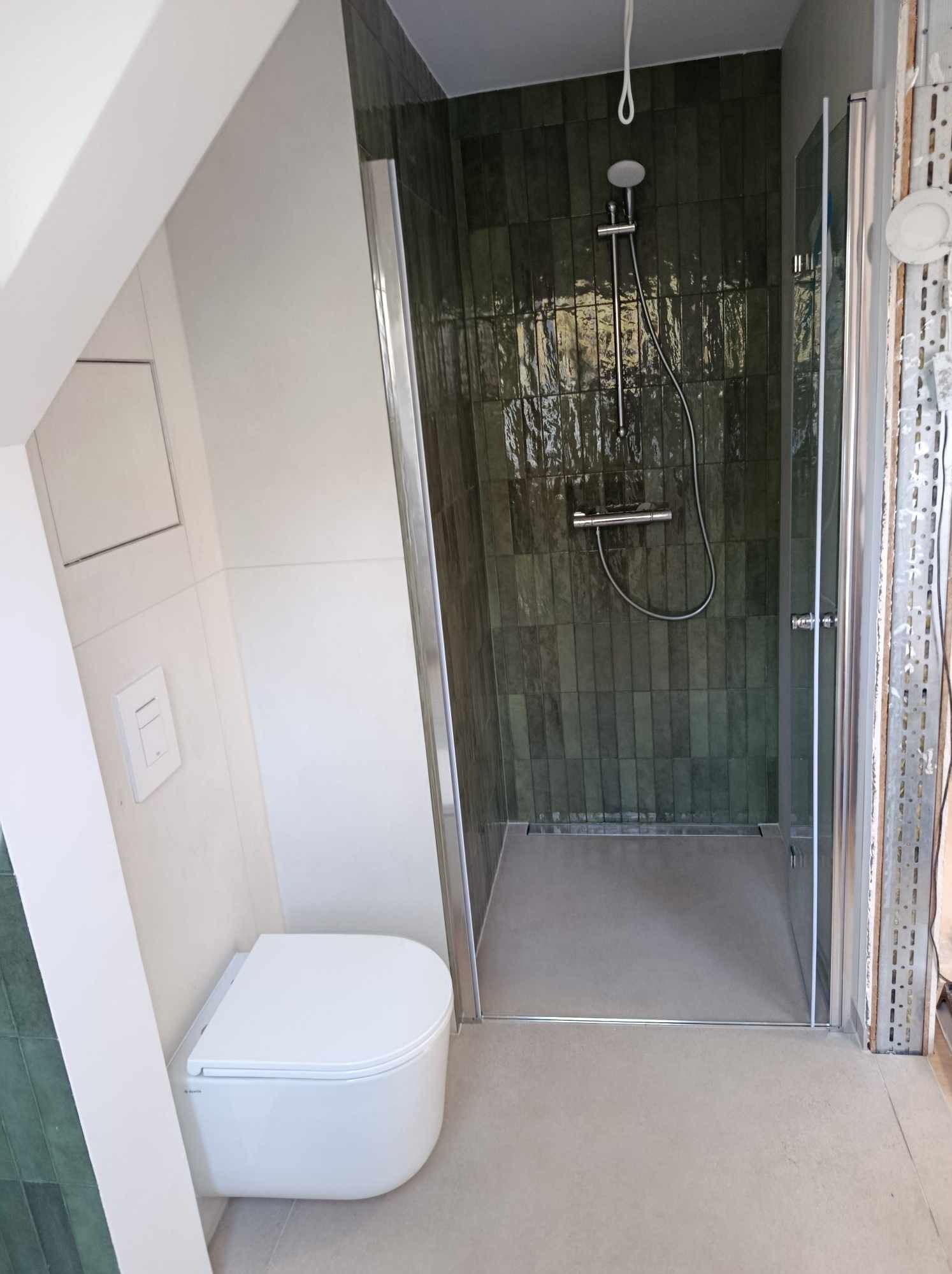 Nowoczesna łazienka w trakcie wykończenia: kabina prysznicowa ze szklanymi drzwiami, ściany wyłożone pionowymi, ciemnozielonymi płytkami z efektem szkliwa, podwieszana toaleta i surowa ściana...