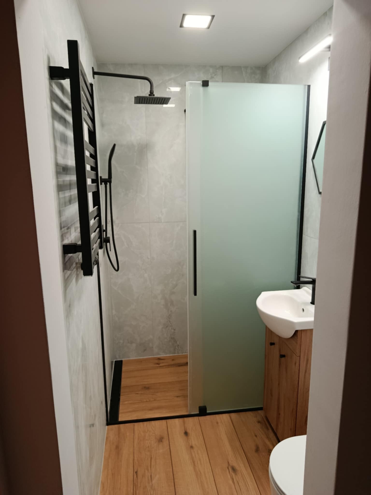 Nowoczesna łazienka po remoncie: kabina prysznicowa ze szkła matowego, drewniana podłoga, umywalka z szafką i czarny grzejnik drabinkowy. Minimalistyczny design.