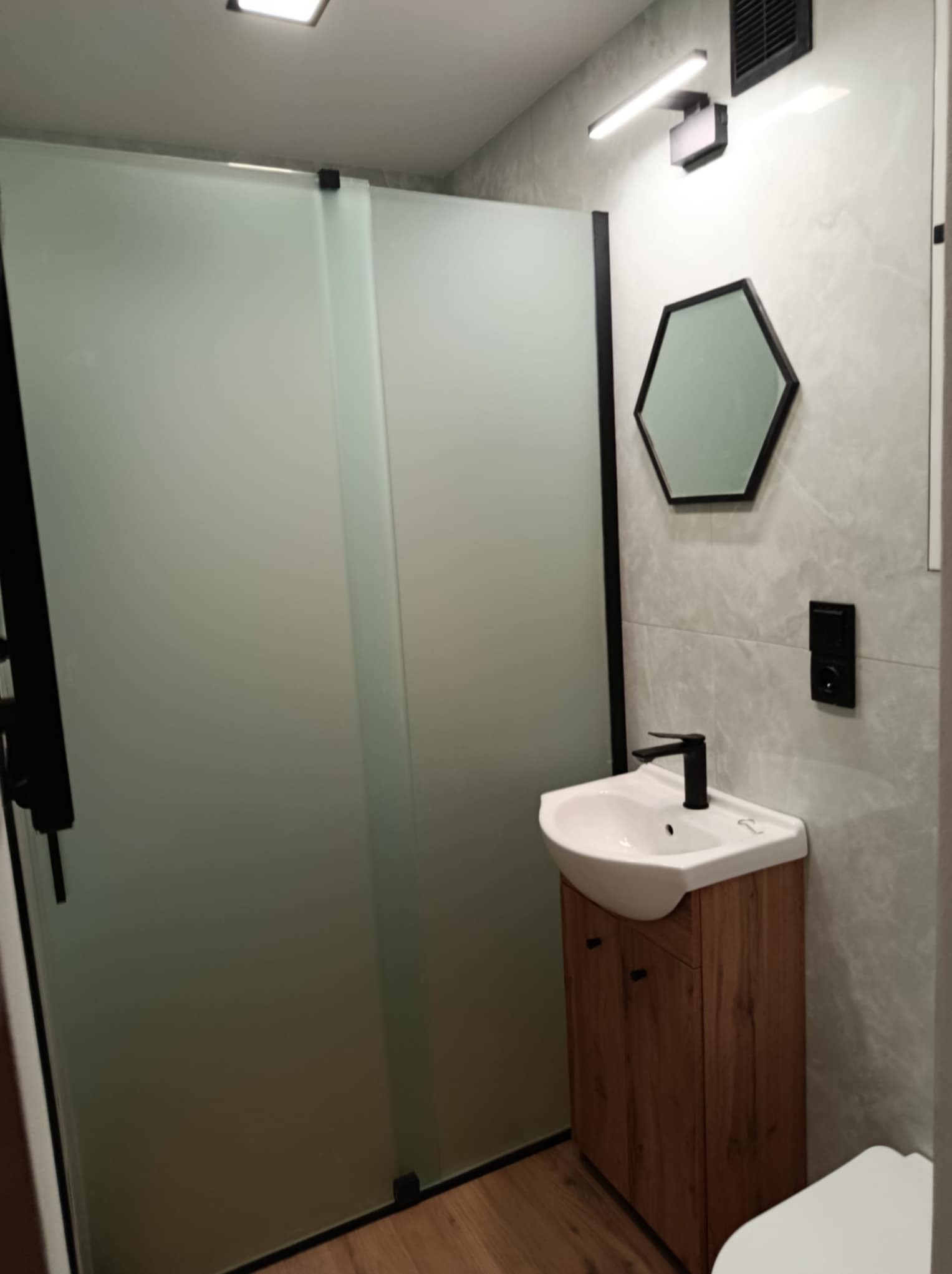 Nowoczesna łazienka z kabiną prysznicową z matowego szkła, heksagonalnym lustrem i szafką pod umywalką z drewnianym fornirem. Minimalistyczny design, szare płytki.