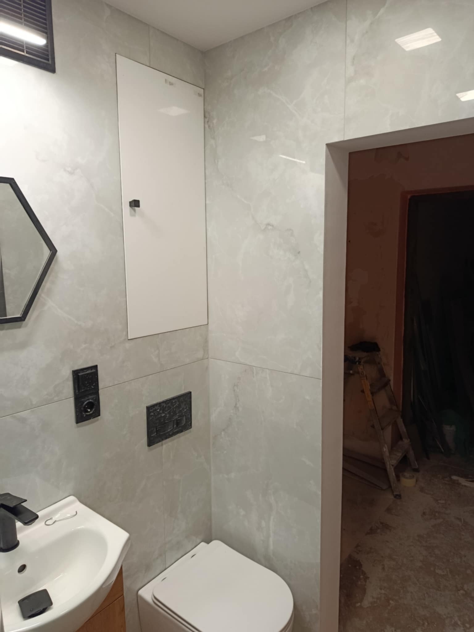 Wykończona łazienka z szarymi płytkami imitującymi marmur, umywalką z szafką, toaletą i lustrem w heksagonalnej ramie. Widoczna klapa rewizyjna i czarne akcenty.