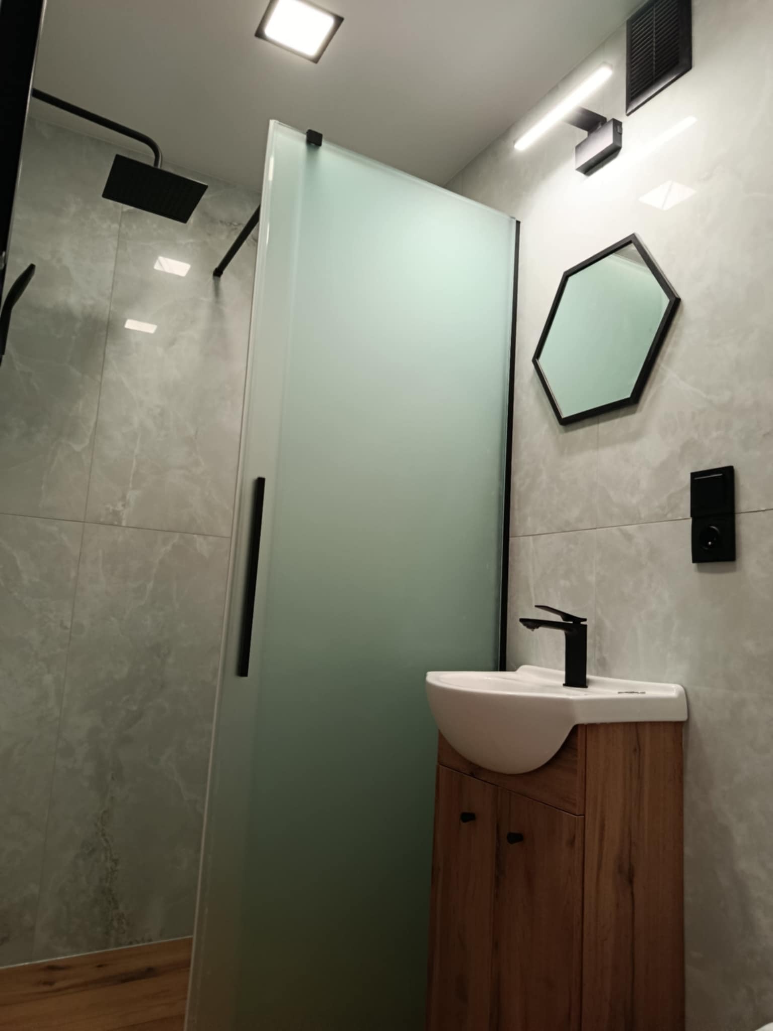 Nowoczesna łazienka z kabiną prysznicową ze szkła, umywalką na drewnianej szafce i heksagonalnym lustrem na tle marmurowych płytek. Czarna armatura i oświetlenie.