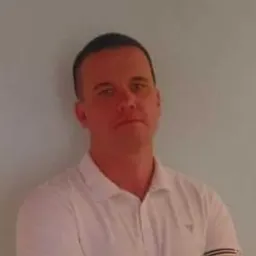 Portret mężczyzny w białej koszulce polo, z założonymi rękami, na tle jasnej, jednolitej ściany. Widoczny cień na ścianie za mężczyzną.