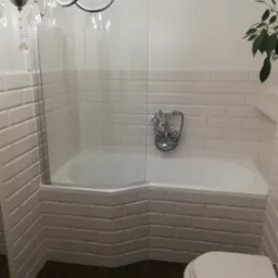 Biała łazienka z wanną obudowaną kafelkami typu metro, szklanym parawanem nawannowym, chromowaną armaturą retro, wiszącym żyrandolem i ciemną podłogą z paneli.