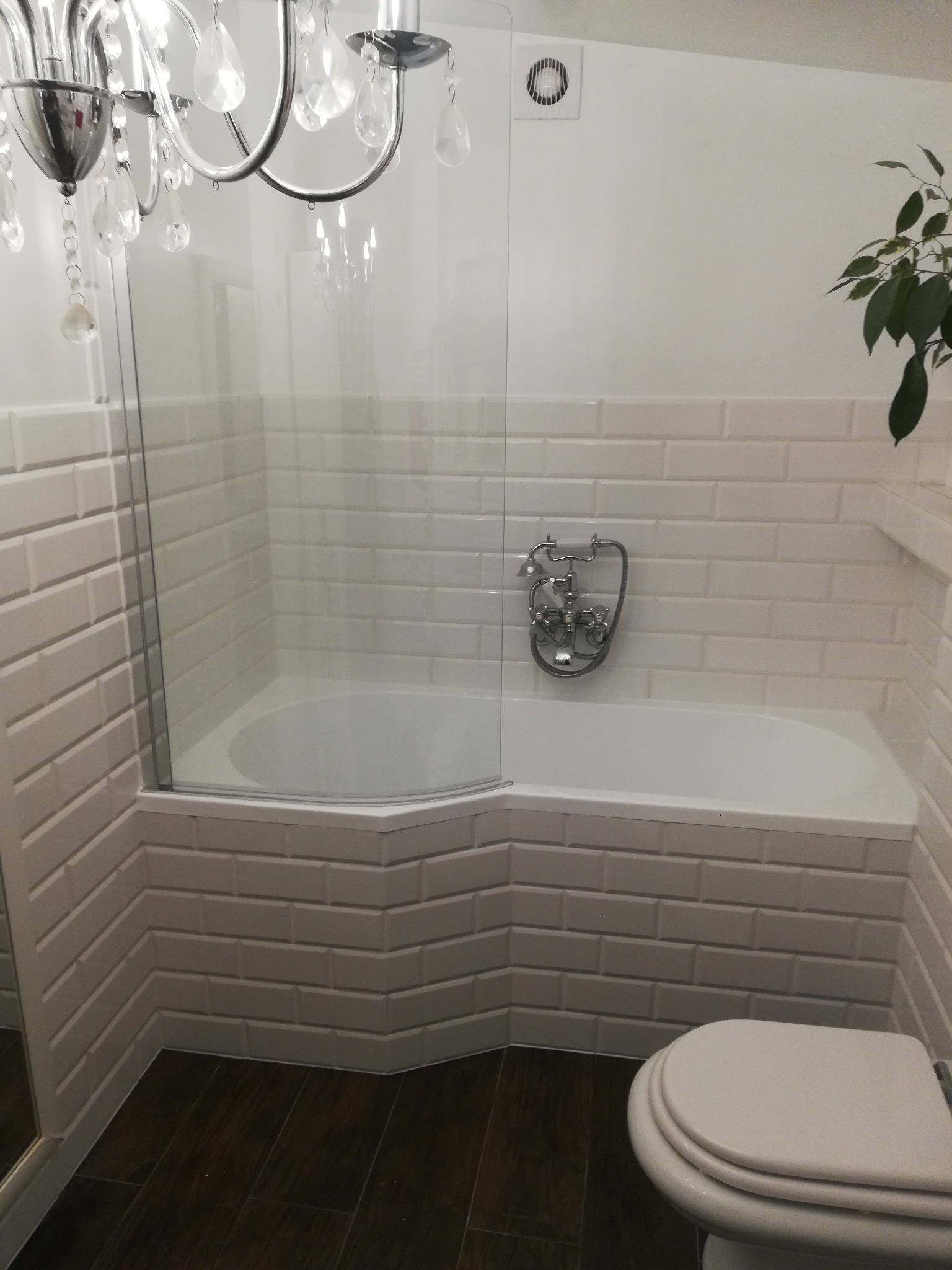 Biała łazienka z wanną obudowaną kafelkami typu metro, szklanym parawanem nawannowym, chromowaną armaturą retro, wiszącym żyrandolem i ciemną podłogą z paneli.