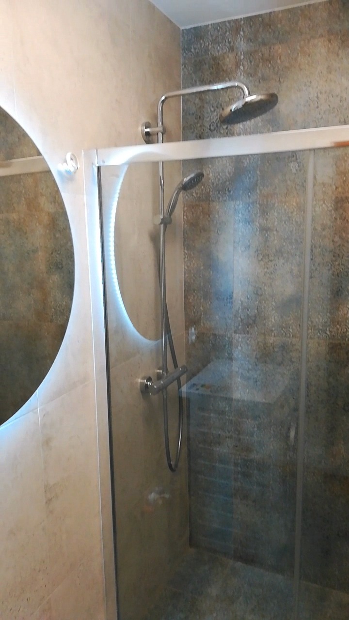 Nowoczesna kabina prysznicowa z deszczownicą i oświetlonym lustrem w łazience wykończonej płytkami imitującymi kamień.