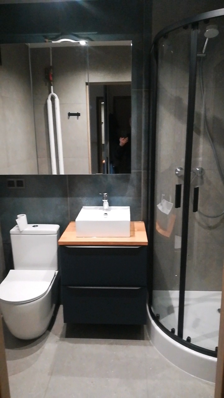 Nowoczesna łazienka z kabiną prysznicową z czarnymi ramami, białą umywalką nablatową na szafce z szufladami w kolorze granatowym, toaletą typu Geberit i dużym lustrem z oświetleniem.