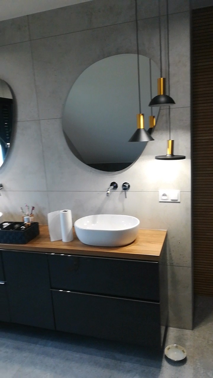 Nowoczesna łazienka z dwoma okrągłymi lustrami, białą umywalką nablatową, drewnianym blatem i czarnymi szafkami, oświetlona trzema wiszącymi lampami z elementami złotymi, z ceramiczną miseczką...