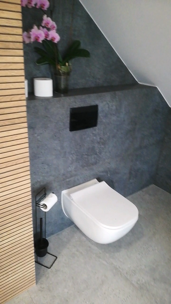 Nowoczesna toaleta podwieszana w łazience ze ścianą wykończoną w szarym kamieniu, drewniane lamele na ścianie po lewej stronie, nad toaletą półka z kwiatami orchidei i rolką papieru toaletowego.