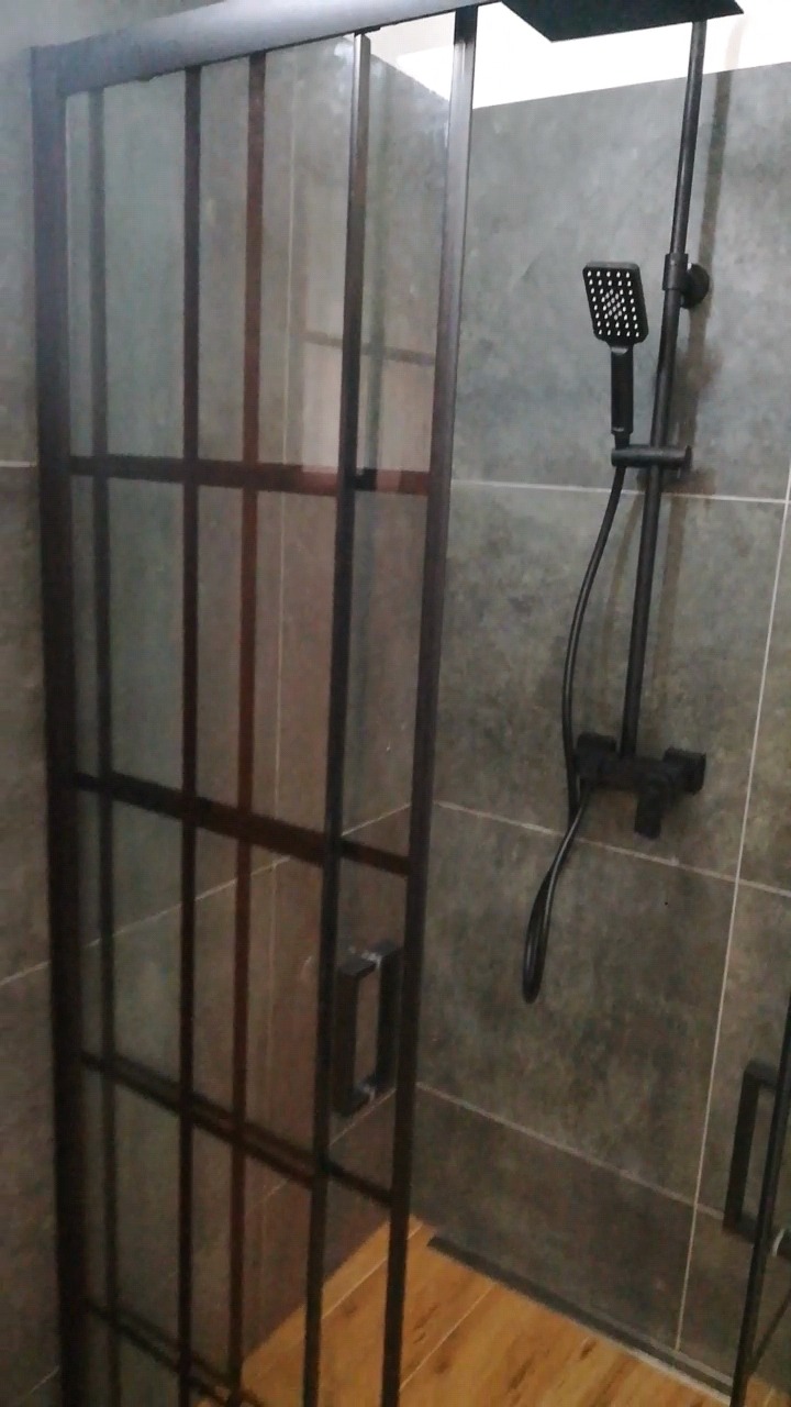 Kabina prysznicowa z czarnymi profilami i szklanymi ściankami, zestaw prysznicowy w kolorze czarnym, szare płytki na ścianach i drewnopodobna podłoga w łazience.