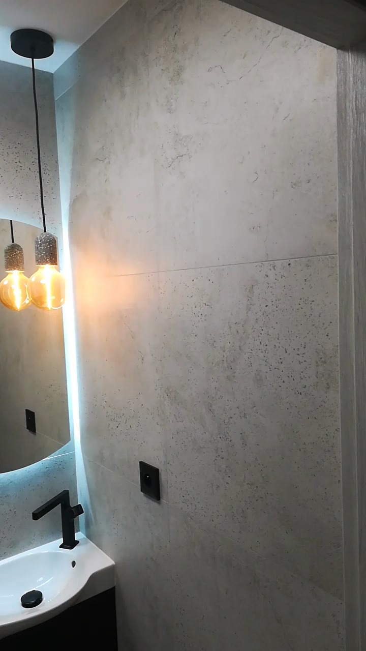 Nowoczesna łazienka z umywalką, czarną armaturą, lustrem z podświetleniem LED i dekoracyjną lampą wiszącą z żarówkami Edisona na tle ściany wykończonej płytkami imitującymi beton.