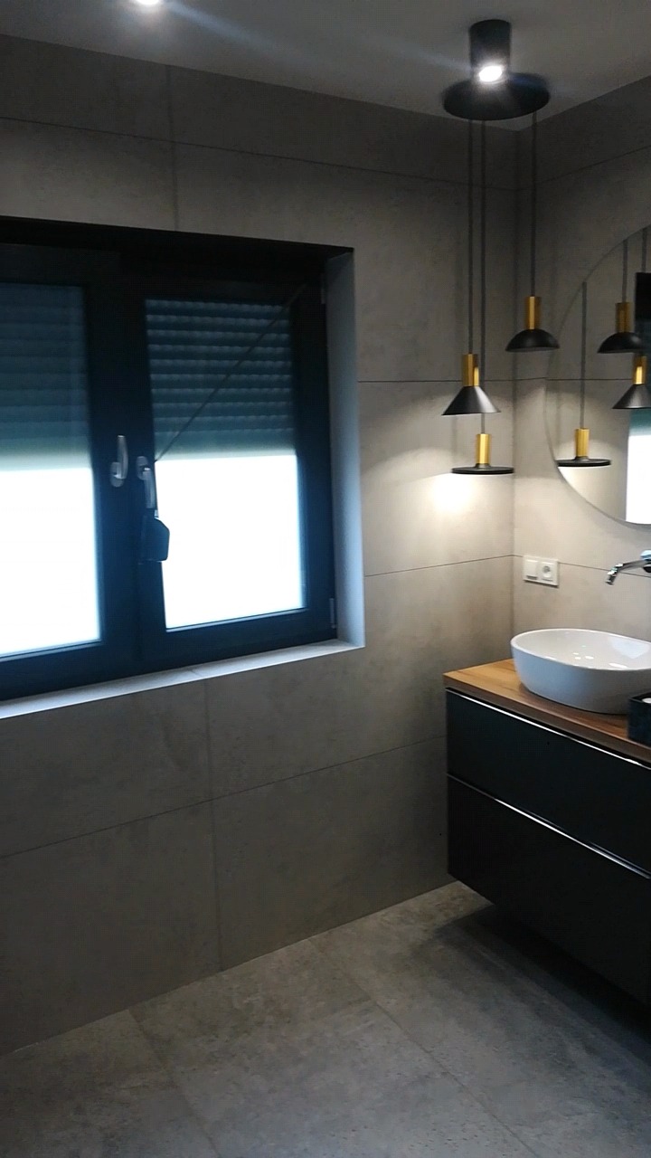 Nowoczesna łazienka z szarymi płytkami, czarną szafką pod umywalką z drewnianym blatem, okrągłym lustrem i designerskimi lampami wiszącymi.