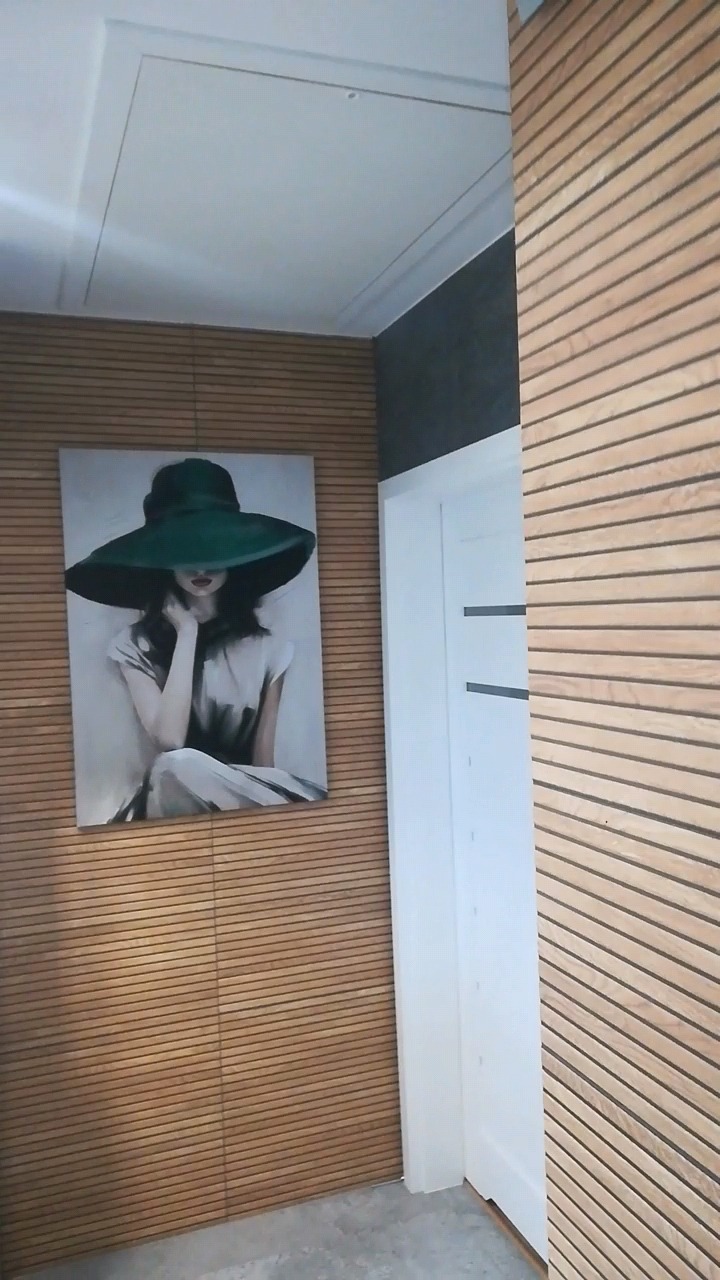 Drewniana ściana z poziomymi listwami, ozdobiona obrazem kobiety w kapeluszu, białe drzwi z poziomymi wstawkami i szara ściana w tle.