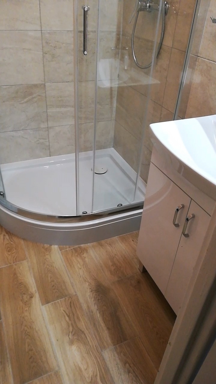 Nowoczesna łazienka po remoncie: kabina prysznicowa z brodzikiem, umywalka z szafką w jasnych kolorach oraz podłoga wyłożona panelami imitującymi drewno.