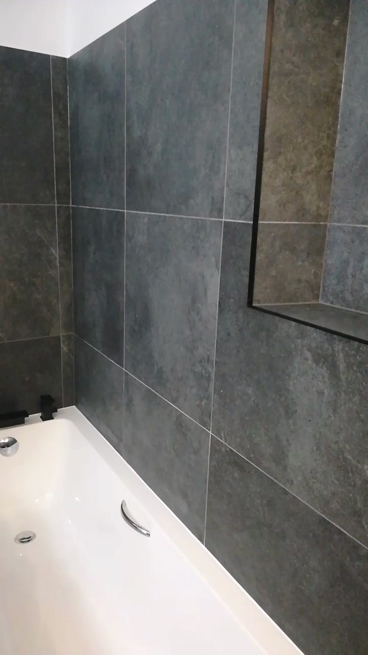 Nowoczesna łazienka: Szare, matowe płytki imitujące kamień na ścianie nad białą wanną z chromowanymi uchwytami i czarną armaturą, wbudowana półka w ścianie.