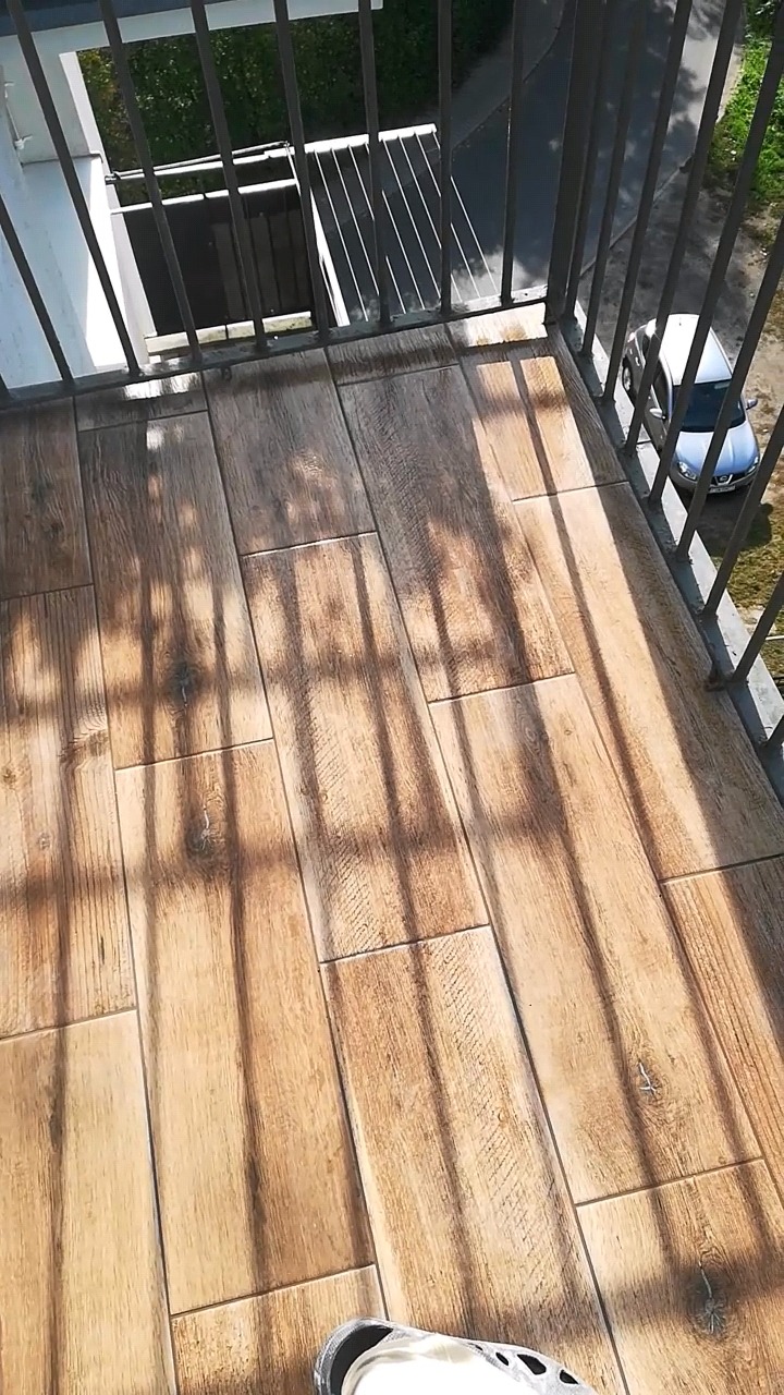 Balkon z nową podłogą z płytek imitujących drewno, widoczne cienie barierki i fragment buta.