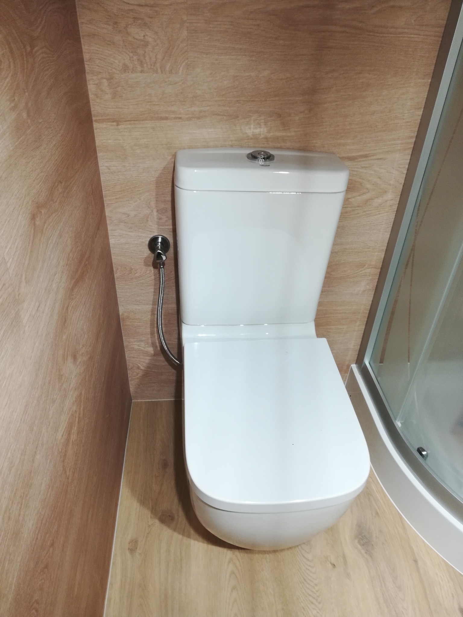 Nowoczesna toaleta z bidetką w łazience wykończonej panelami imitującymi drewno, widoczny fragment kabiny prysznicowej.