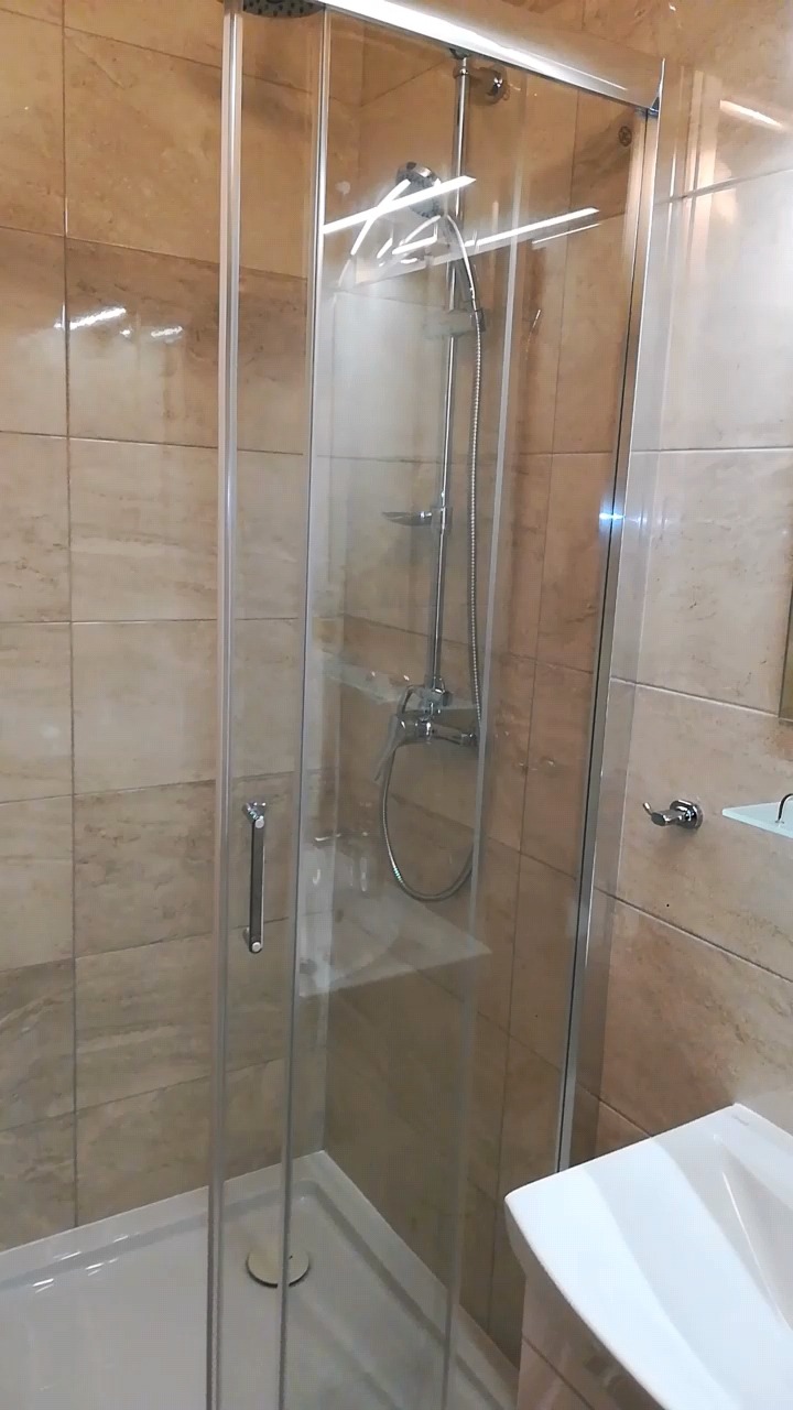 Szklana kabina prysznicowa z chromowaną armaturą, jasnobeżowe płytki na ścianach, biały brodzik i umywalka.