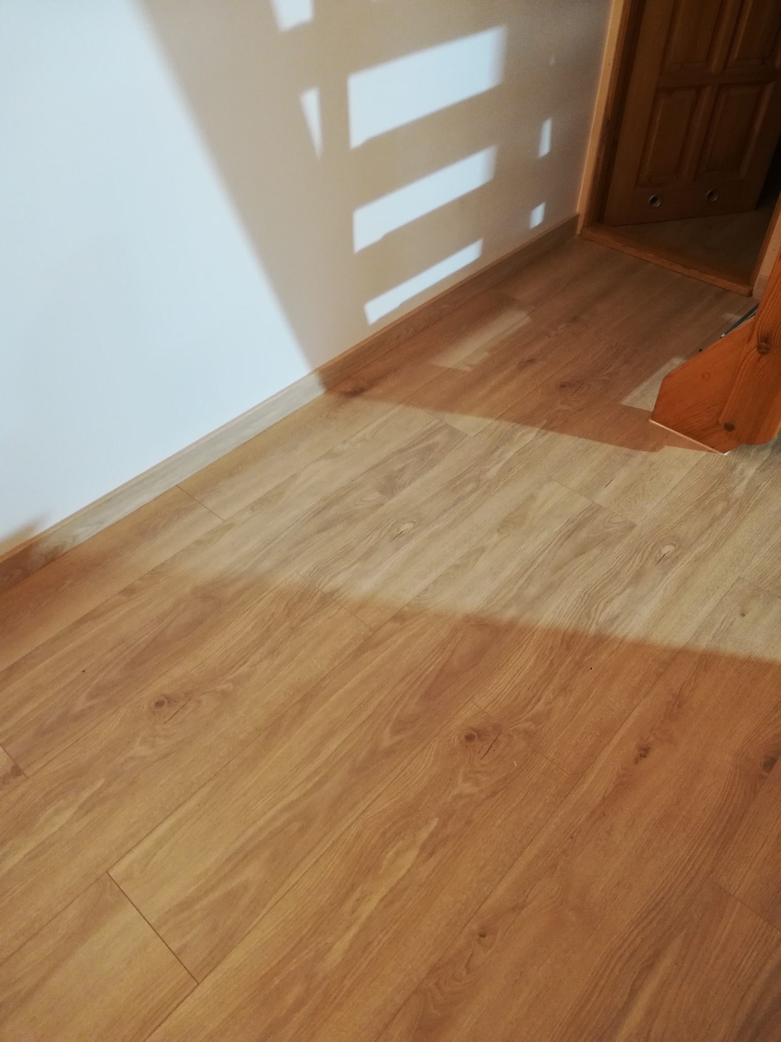 Ułożone panele podłogowe o wzorze drewna, widoczne przy ścianie z listwą przypodłogową i fragment drzwi, oświetlone przez padające światło z zewnątrz.