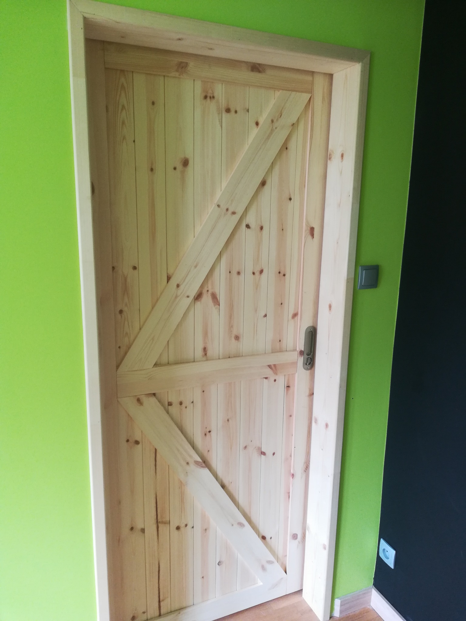 Surowe drewniane drzwi w stylu rustykalnym z ukośnymi wzmocnieniami na tle zielonej i czarnej ściany, widoczny fragment podłogi i włącznik światła.