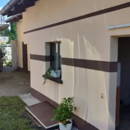 Elewacja domu po malowaniu z poziomymi pasami w kolorze brązowym, doniczka z rośliną przy wejściu, roślina w doniczce na parapecie okna.