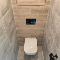 Wąska toaleta z nowoczesną białą miską WC z elektronicznym panelem, wykończona płytkami imitującymi drewno na górnej części ściany i szarymi płytkami na dole, uzupełniona czarnym przyciskiem...