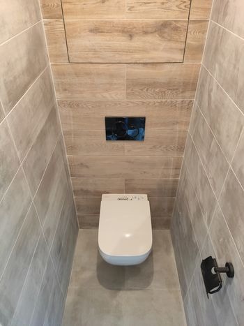 Wąska toaleta z nowoczesną białą miską WC z elektronicznym panelem, wykończona płytkami imitującymi drewno na górnej części ściany i szarymi płytkami na dole, uzupełniona czarnym przyciskiem...
