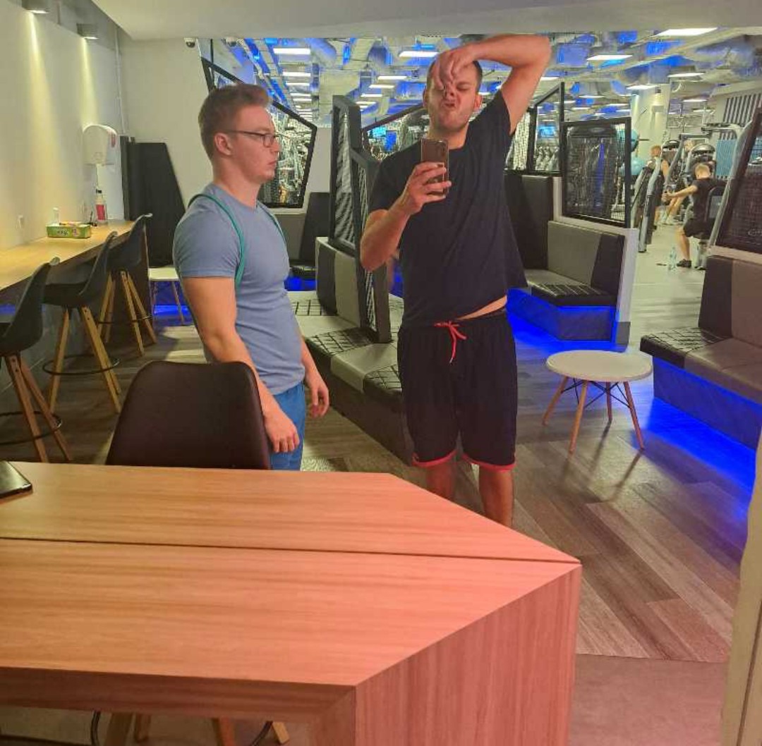 Dwaj mężczyźni w sportowych strojach robią selfie w siłowni, widoczne ławki i sprzęt do ćwiczeń w tle.