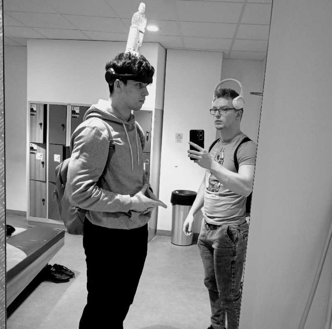 Mężczyzna w szarej bluzie z kapturem i butelką na głowie pozuje do zdjęcia robionego w lustrze przez drugiego mężczyznę w okularach i słuchawkach, na tle szatni z szafkami.