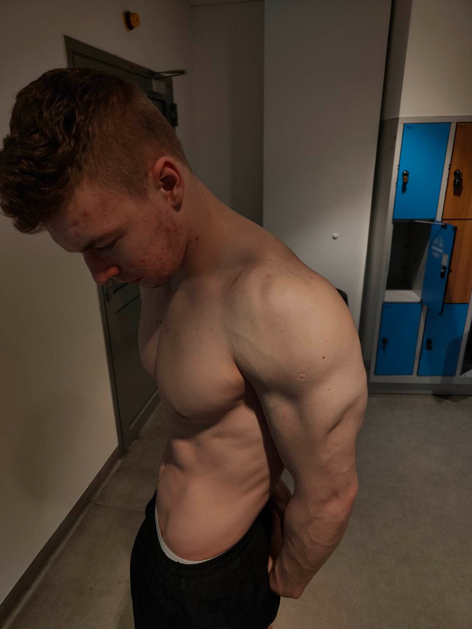 Mężczyzna z widoczną muskulaturą pozuje w szatni, z boku widoczne niebiesko-brązowe szafki.