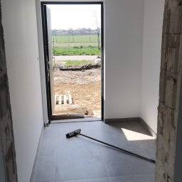 MarSop Mariusz Sopel - Wnętrze w trakcie wykańczania: świeżo pomalowane ściany, szare płytki podłogowe, widoczny fragment surowego muru i widok na zewnątrz przez otwarte drzwi.