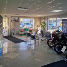 Jasne wnętrze serwisu motocyklowego z trzema motocyklami na pierwszym planie, stolik z krzesłami dla klientów w tle, reklama Castrol oraz beczki Castrol jako dekoracja.