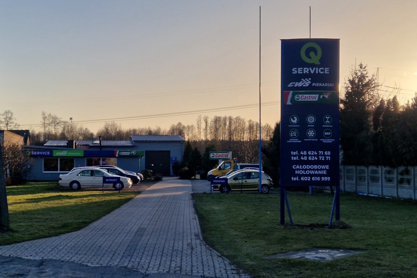 Budynek serwisu samochodowego z logo firmy i reklamą olejów Castrol, widoczne zaparkowane auta i tablica informacyjna z numerami telefonów.