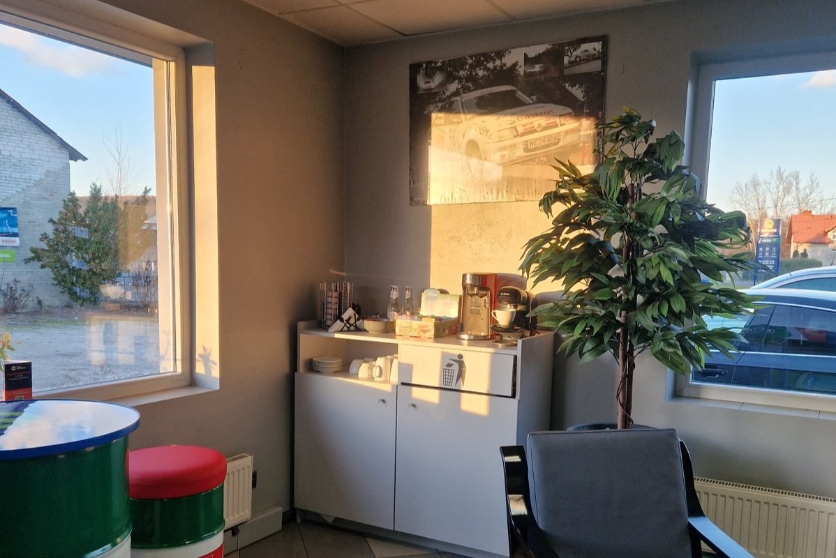 Poczekalnia w warsztacie samochodowym: stolik z beczki, automat do kawy, dekoracja w postaci plakatu rajdowego samochodu i sztuczna roślina w rogu.