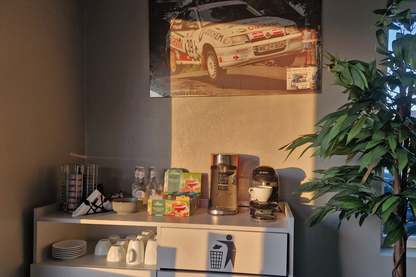 Poczekalnia warsztatu samochodowego: plakat rajdowego Opla Astra GSI z numerem 39 obok ekspresu do kawy i sztucznego drzewka.