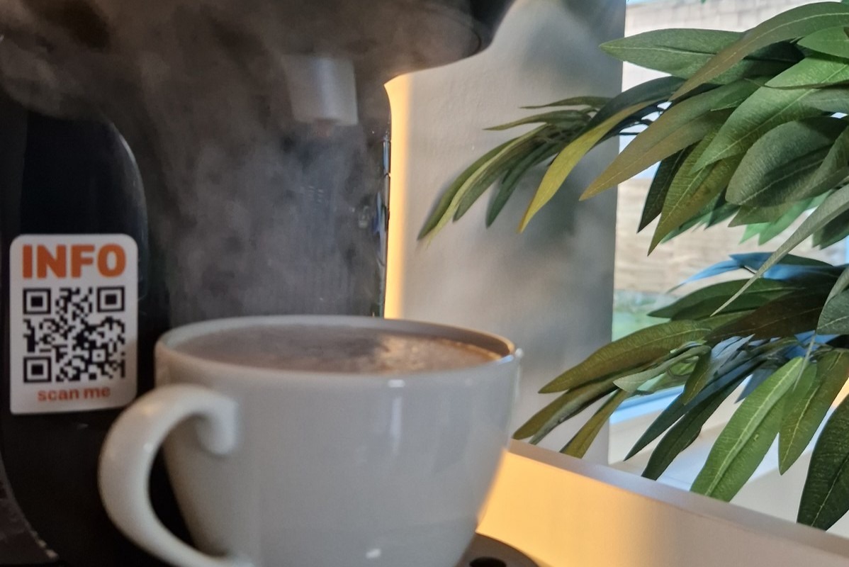 Ekspres do kawy w trakcie pracy, para unosi się nad filiżanką, obok naklejka z kodem QR i rośliną doniczkową w tle