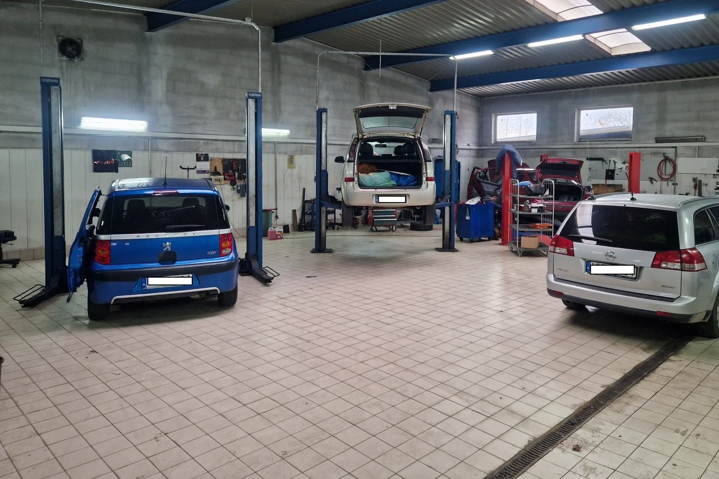 Wnętrze warsztatu samochodowego z trzema samochodami: niebieski Peugeot 1007 z otwartymi drzwiami, biały samochód na podnośniku z otwartą klapą bagażnika i srebrny Opel Vectra kombi. Widoczne...