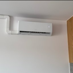 ASKLIM Alan Stencel - Klimatyzator marki Gree zamontowany na ścianie w pomieszczeniu w Przodkowie, z widocznymi białymi rurami instalacyjnymi i fragmentem drewnianej zabudowy meblowej.