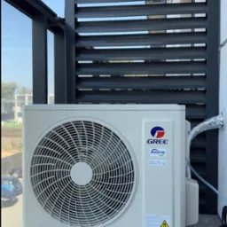 Zewnętrzna jednostka klimatyzacji marki Gree zamontowana na balkonie z szarymi żaluzjami, widoczna srebrna rura odprowadzająca skropliny.