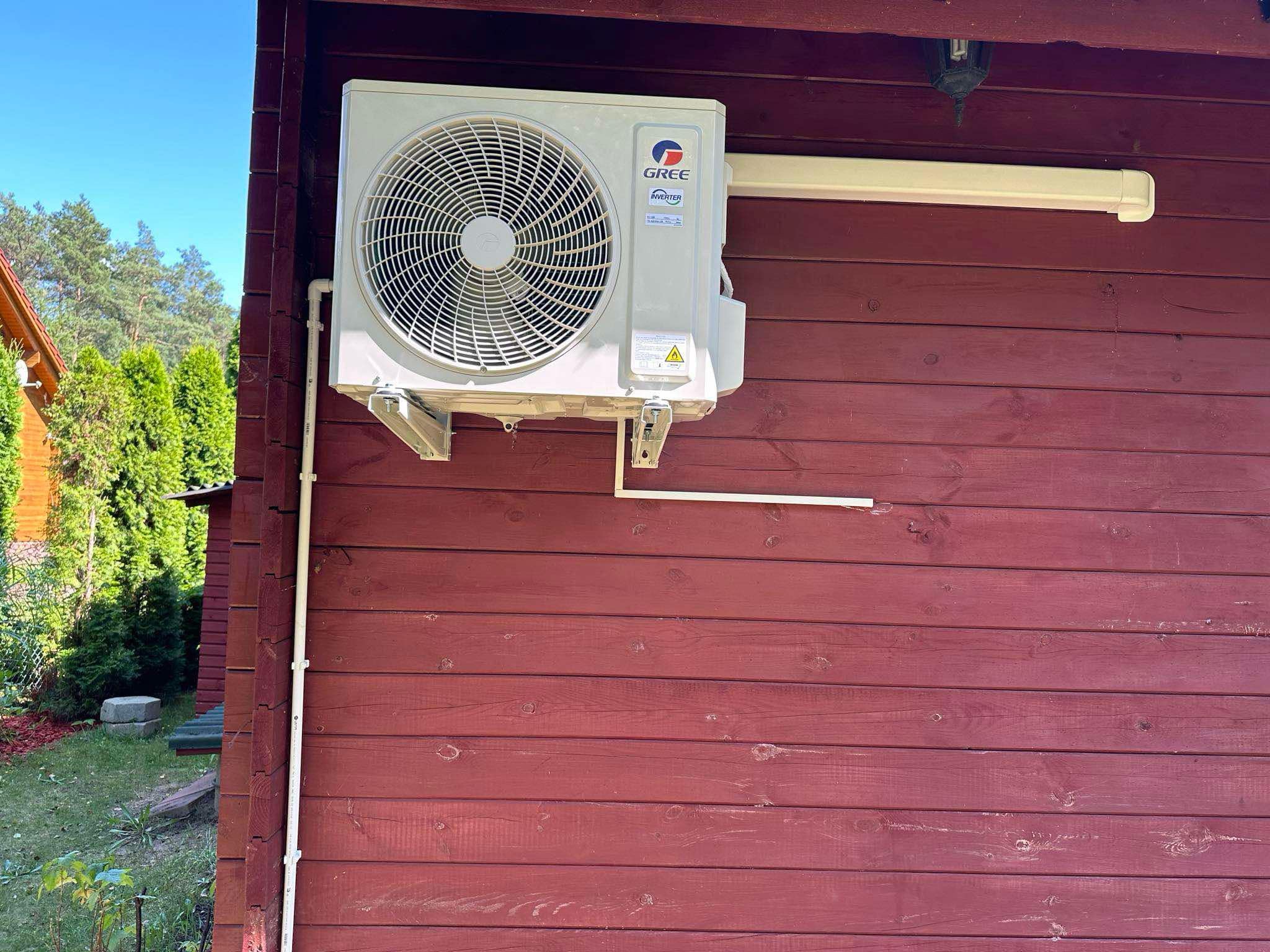 Zewnętrzna jednostka klimatyzatora marki Gree zamontowana na ścianie drewnianego domu pomalowanego na czerwono, z widocznymi przewodami i elementami instalacji.