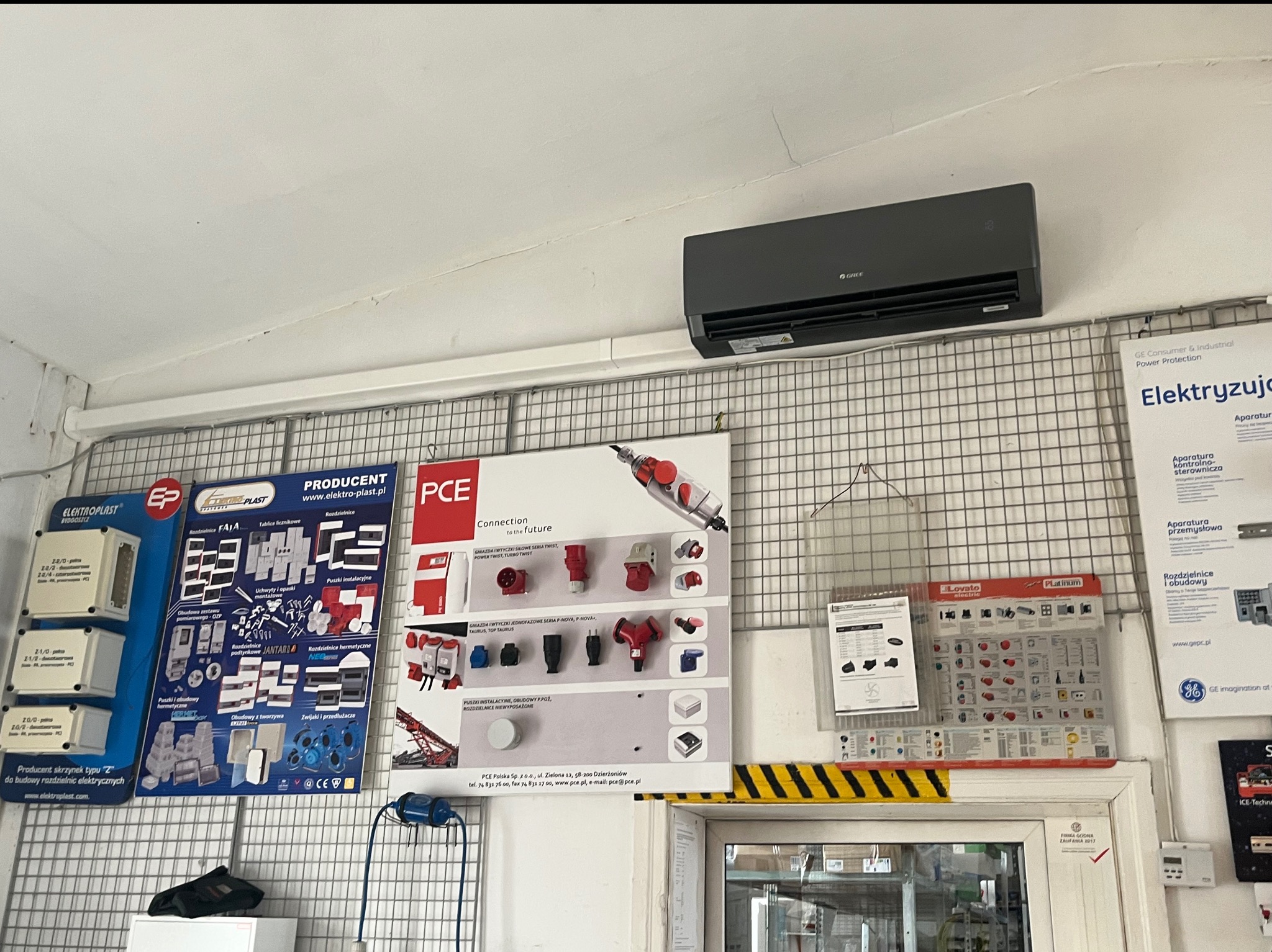 Szara jednostka klimatyzacji marki Gree zamontowana na ścianie nad tablicami informacyjnymi z elementami elektrycznymi i instalacyjnymi w pomieszczeniu sklepowym lub magazynowym.