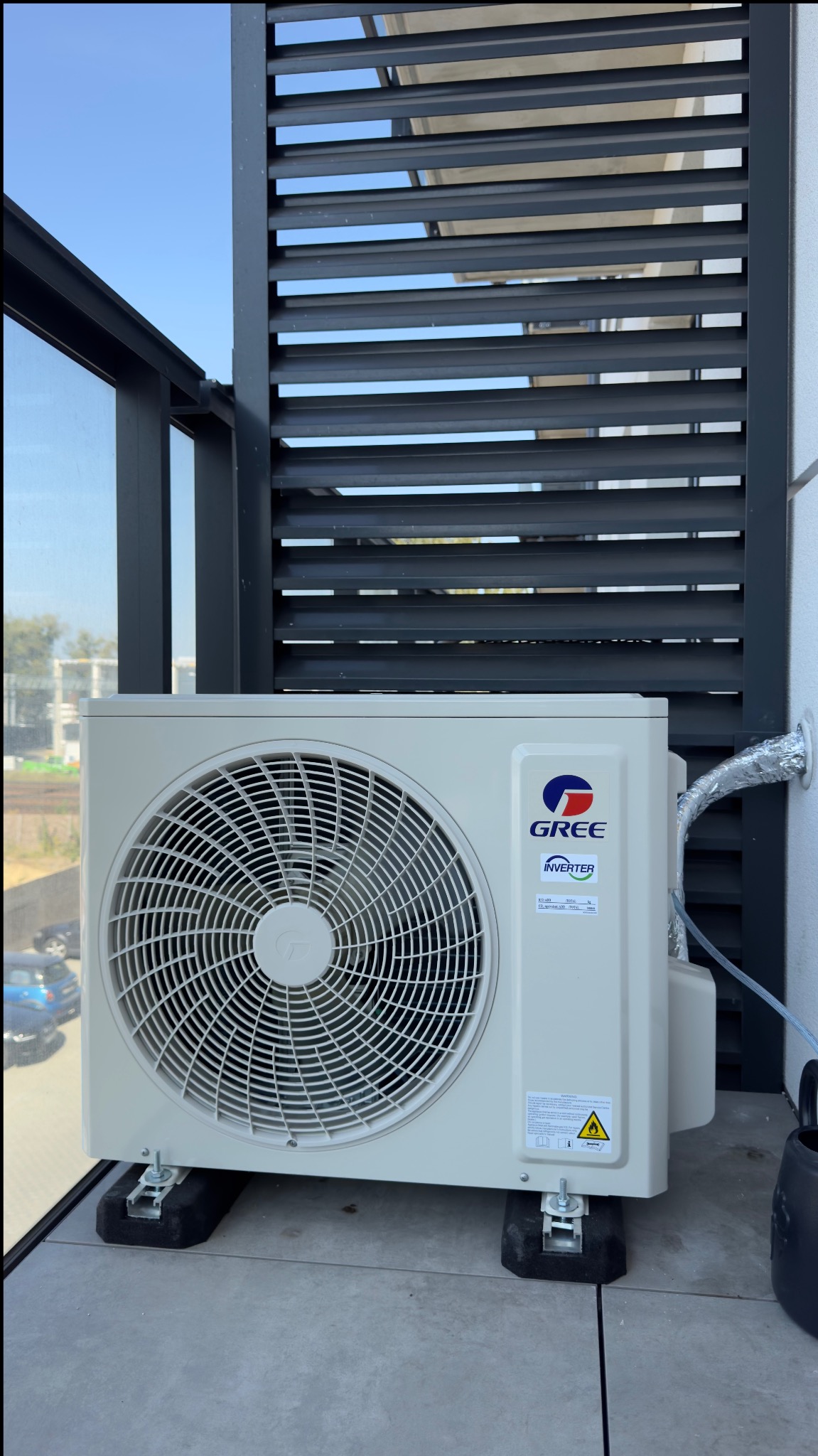 Zewnętrzna jednostka klimatyzacji marki Gree zamontowana na balkonie z szarymi żaluzjami, widoczna srebrna rura odprowadzająca skropliny.