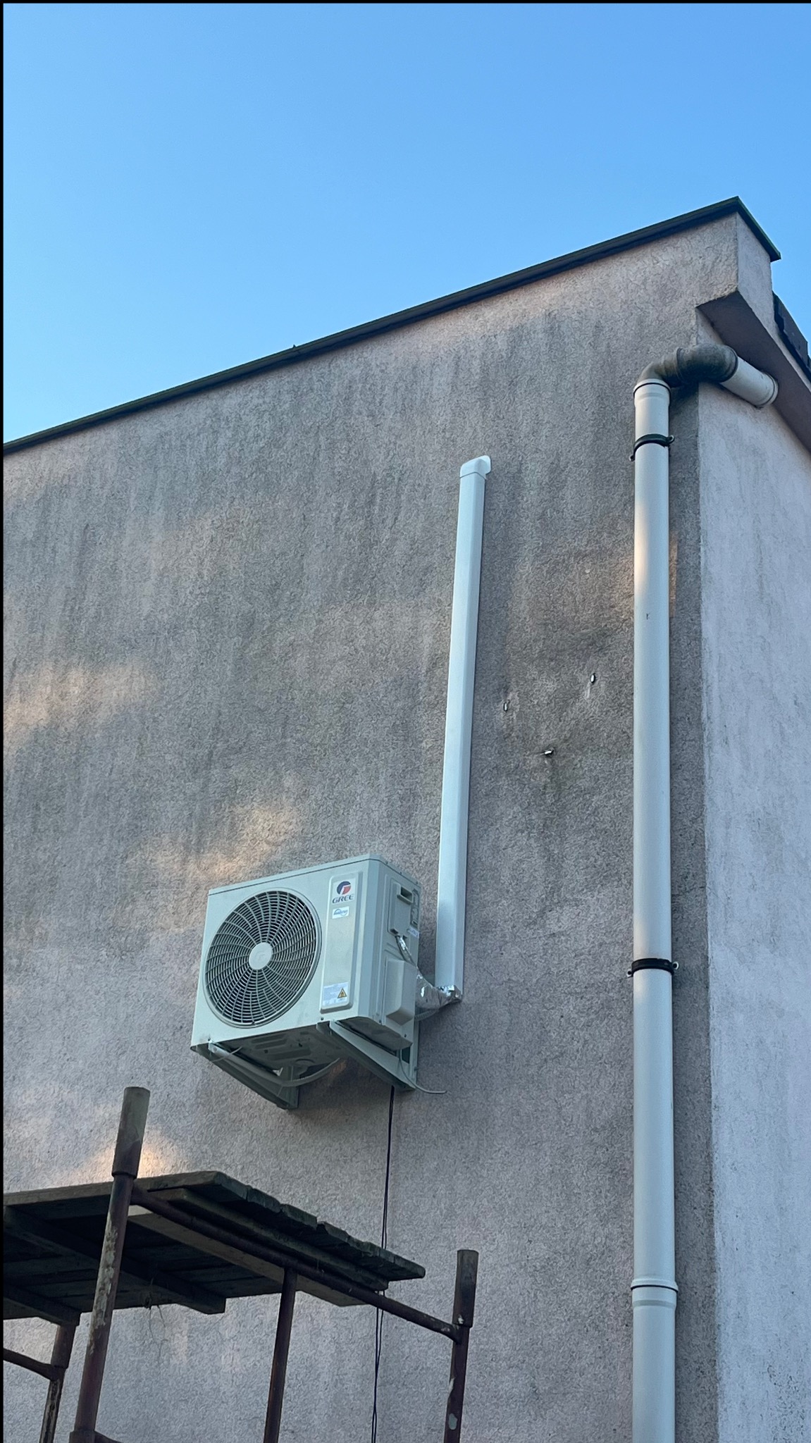 Zewnętrzna jednostka klimatyzatora marki Gree zamontowana na szarej elewacji budynku, widoczne rury odprowadzające skropliny, rusztowanie w dolnej części kadru.