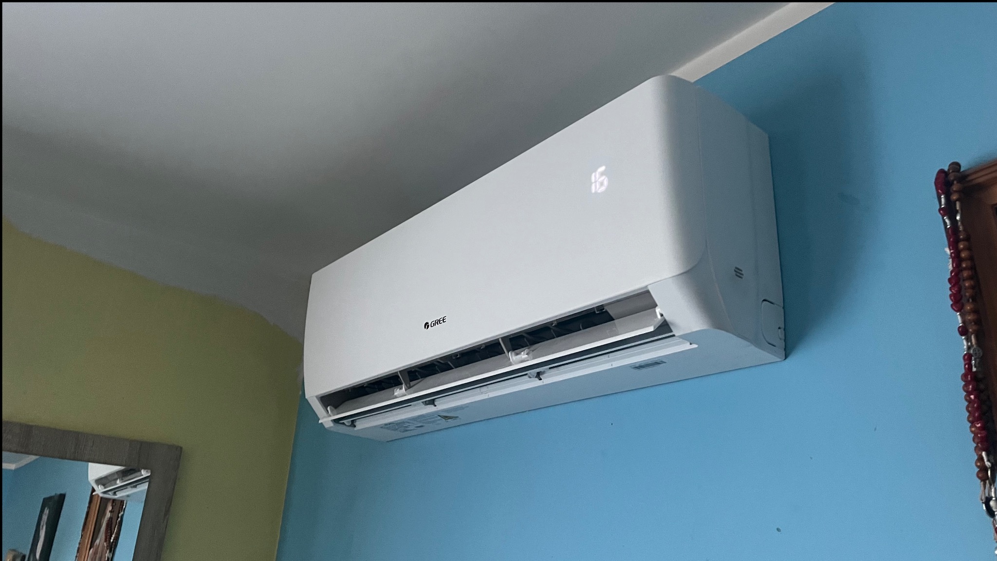 Biała jednostka wewnętrzna klimatyzatora marki Gree zamontowana na błękitnej ścianie w pomieszczeniu o żółto-szarym suficie, z widocznym wyświetlaczem temperatury ustawionym na 16 stopni Celsjusza.