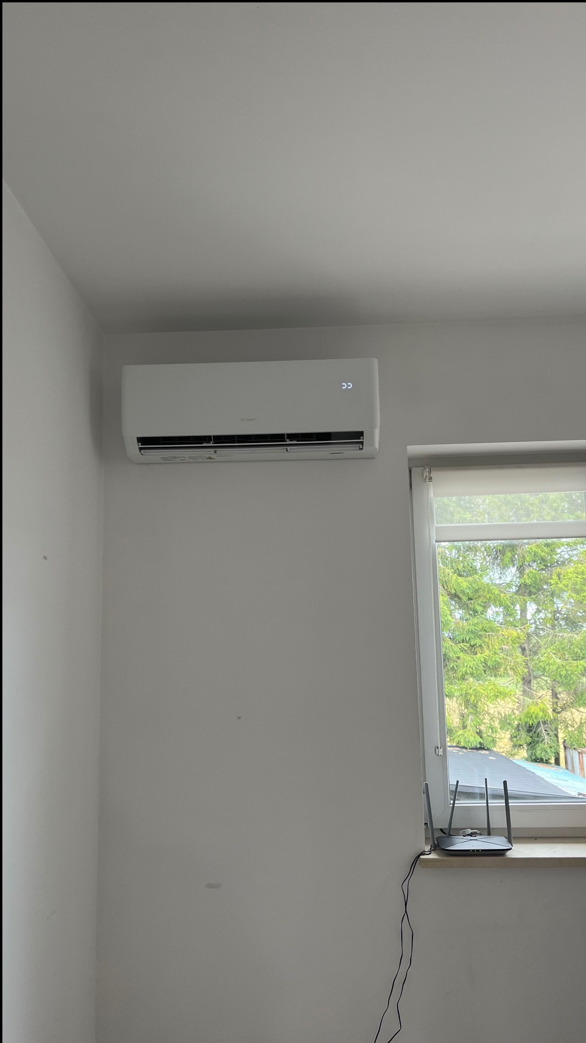Biała jednostka wewnętrzna klimatyzacji zamontowana na ścianie w pomieszczeniu z oknem, obok okna router Wi-Fi.