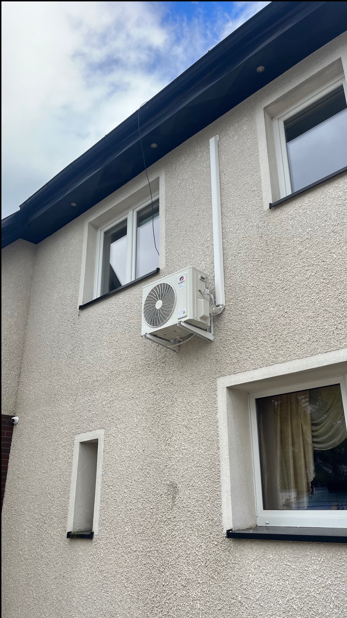 Zewnętrzna jednostka klimatyzacyjna marki Gree zamontowana na elewacji budynku mieszkalnego, widoczne okna z białymi ramami i rura odprowadzająca skropliny.