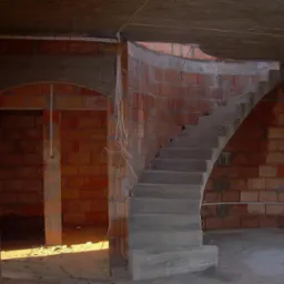 Wnętrze w trakcie budowy: surowe ceglane ściany, betonowe schody łukowe, widoczne instalacje elektryczne, w tle otwór drzwiowy z łukowym nadprożem.